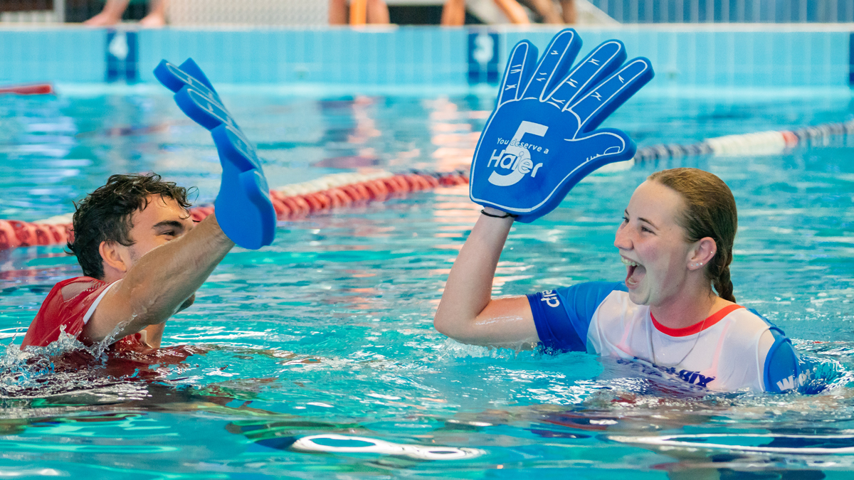Young Man and woman high-fiving in pool with blue Haier foam hands.